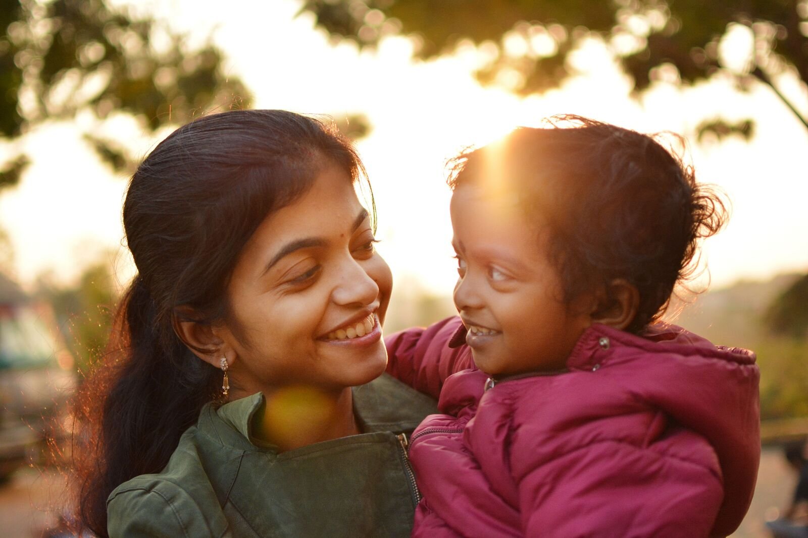 A smiling mother holds her toddler close outdoors in warm evening light
